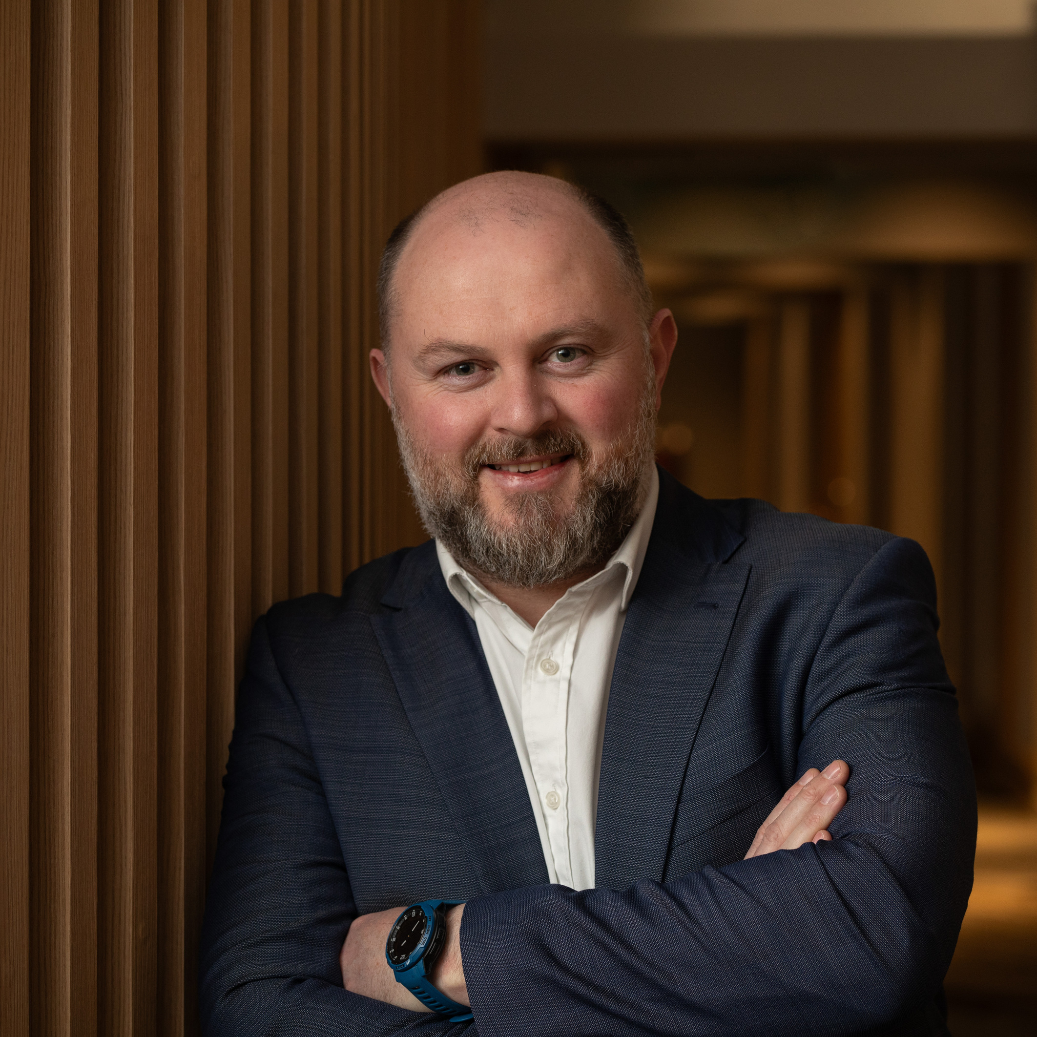 Neil Buggy, in a navy jacket with his arms folded, photographed against a dark timber-slatted wall.