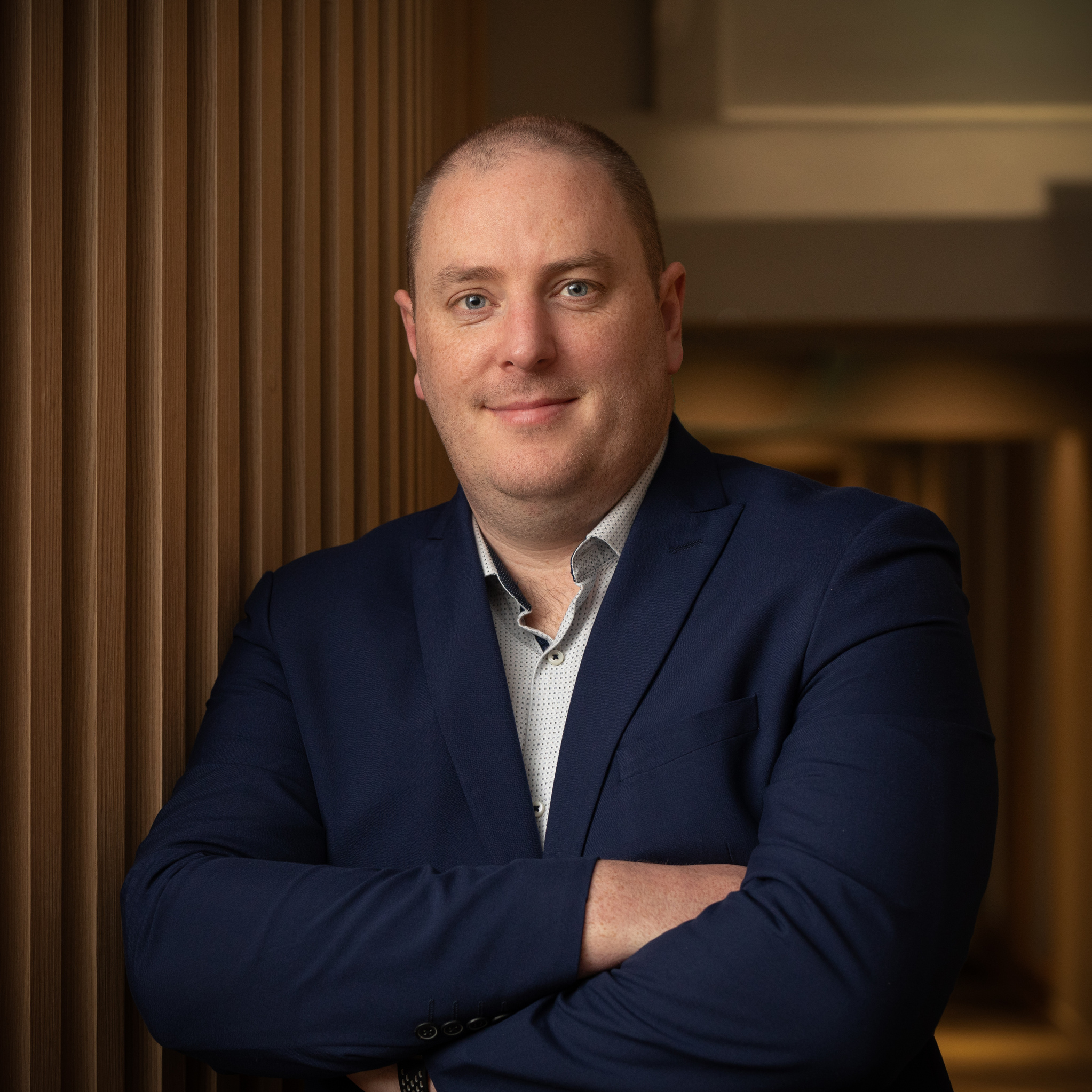 Seamus O'Dwyer, in a navy suit with his arms folded, photographed against the same dark timber-slatted wall.
